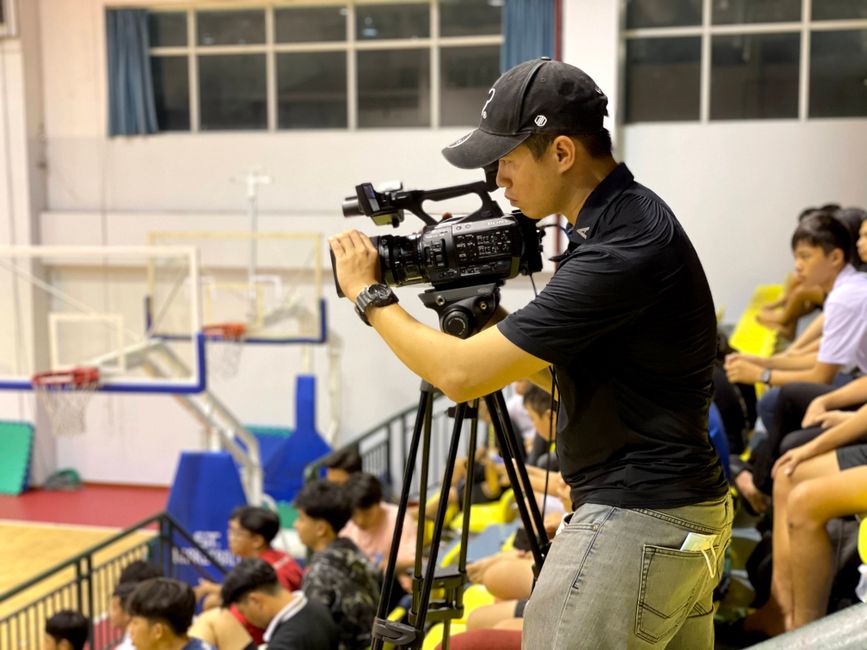 Cameraman capturing a basketball game indoors, with spectators in the background.